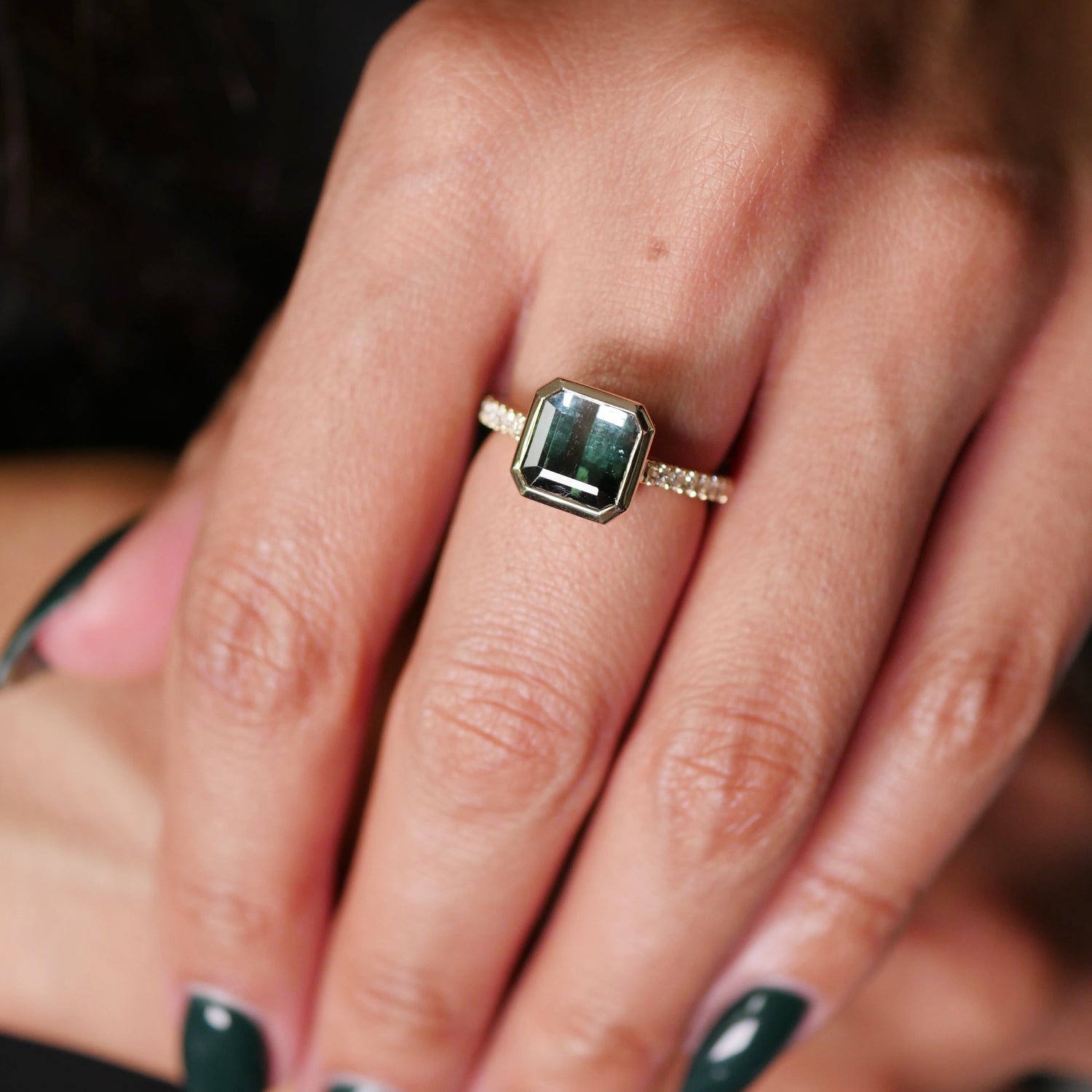 Asscher cut green tourmaline bezel ring in yellow gold with a diamond pavé band, worn on the hand.