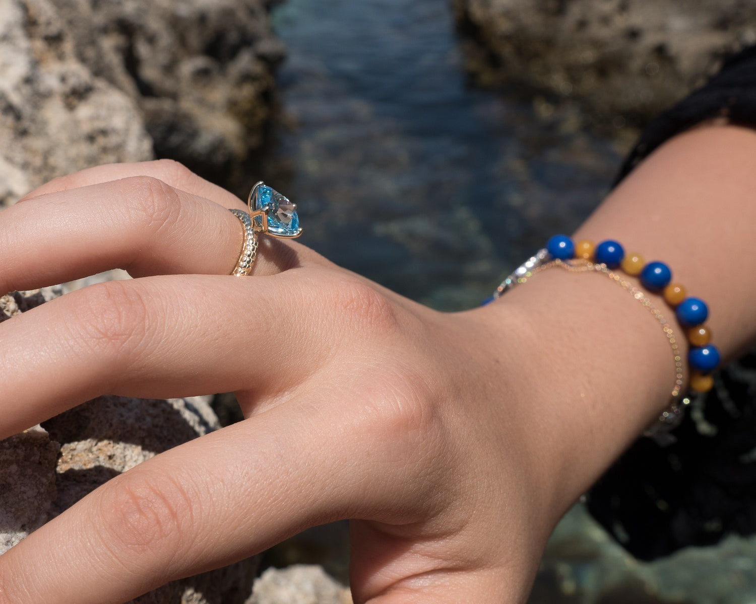Woman wearing a blue topaz ring and a blue and yellow beaded bracelet by Jewelry Design House beside seaside rocks.