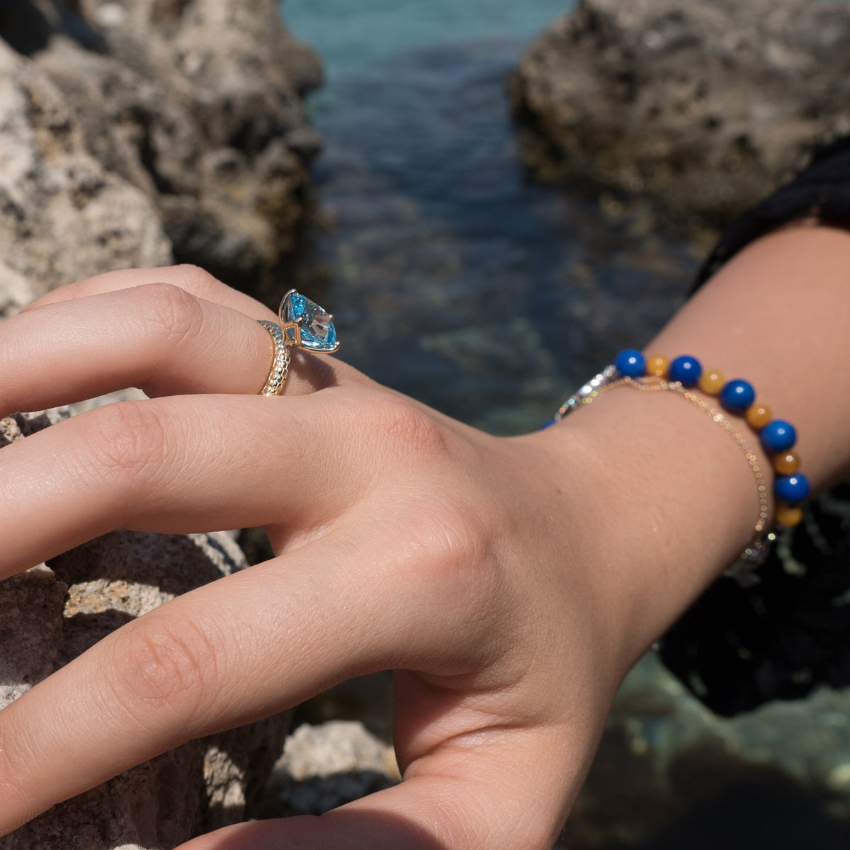 Woman wearing a blue topaz ring and a blue and yellow beaded bracelet by Jewelry Design House beside seaside rocks.