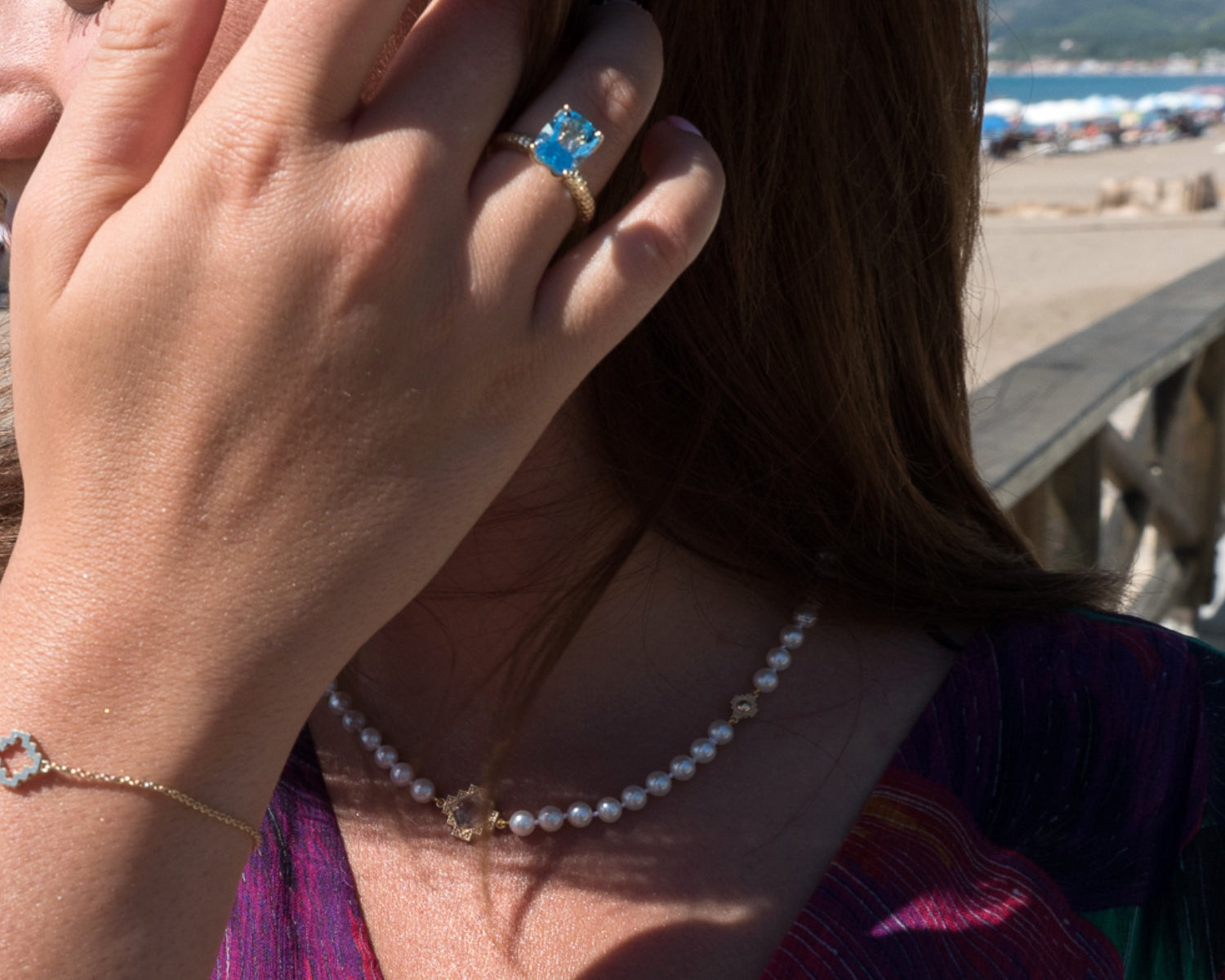 Woman wearing a blue topaz ring, a pearl necklace, and a delicate chain bracelet by Jewelry Design House beside the sea.