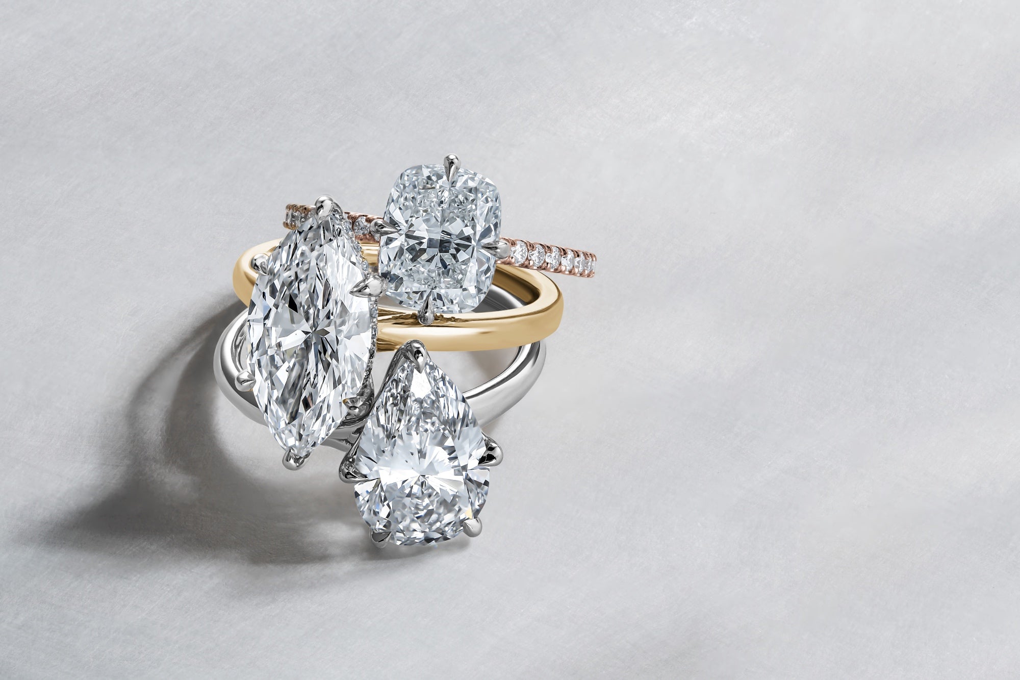 Three diamond engagement rings with marquise, cushion, and pear-shaped centre stones arranged on a light grey background.