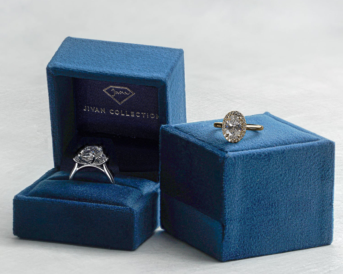 Two diamond rings displayed in and on blue Jivan Collection jewelry boxes against a light grey background.