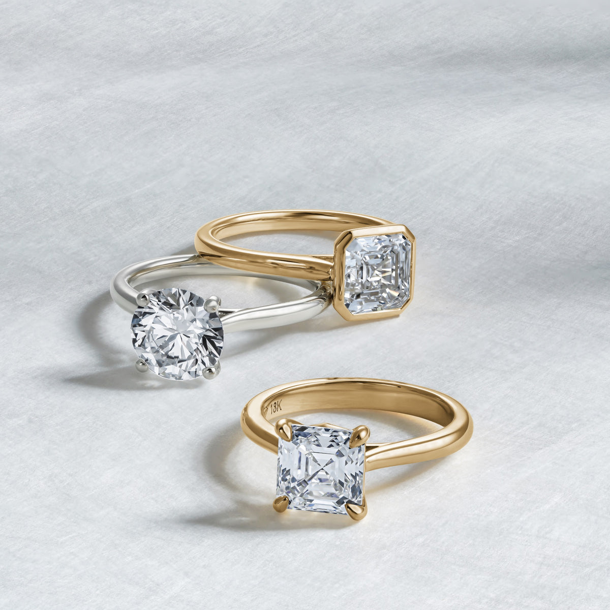 Three diamond solitaire engagement rings in yellow and white gold arranged on grey background.
