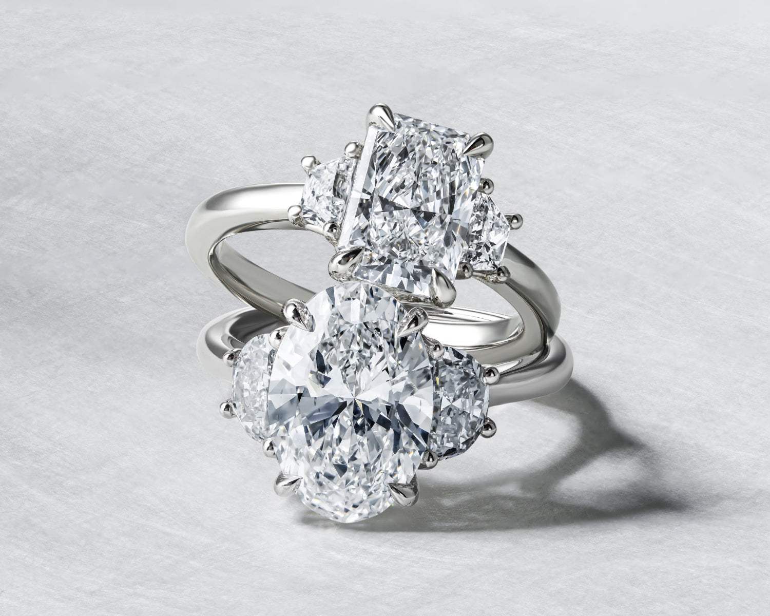 Two three-stone diamond rings with elongated centre stones arranged together on a textured light grey background.