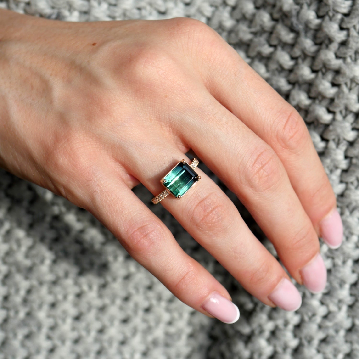 Emerald-cut blue tourmaline east-west ring with a diamond pavé band in 14K yellow gold, worn on a hand.