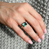 Emerald-cut blue tourmaline east-west ring with a diamond pavé band in 14K yellow gold, worn on a hand.