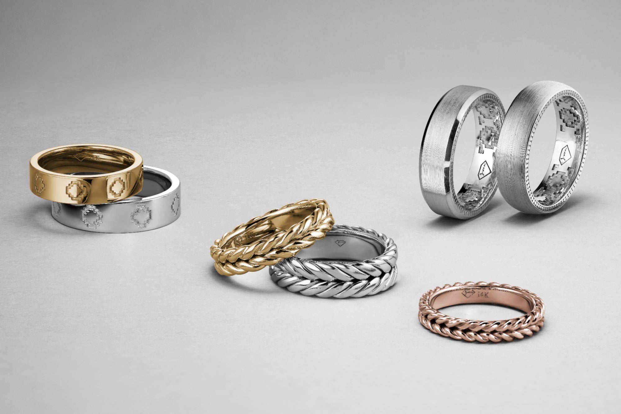 Men’s wedding bands with engraved, braided, and brushed finishes arranged on a light grey background.