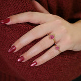 Gold ring with a ruby-studded pomegranate motif, worn on the hand against a red textured background.