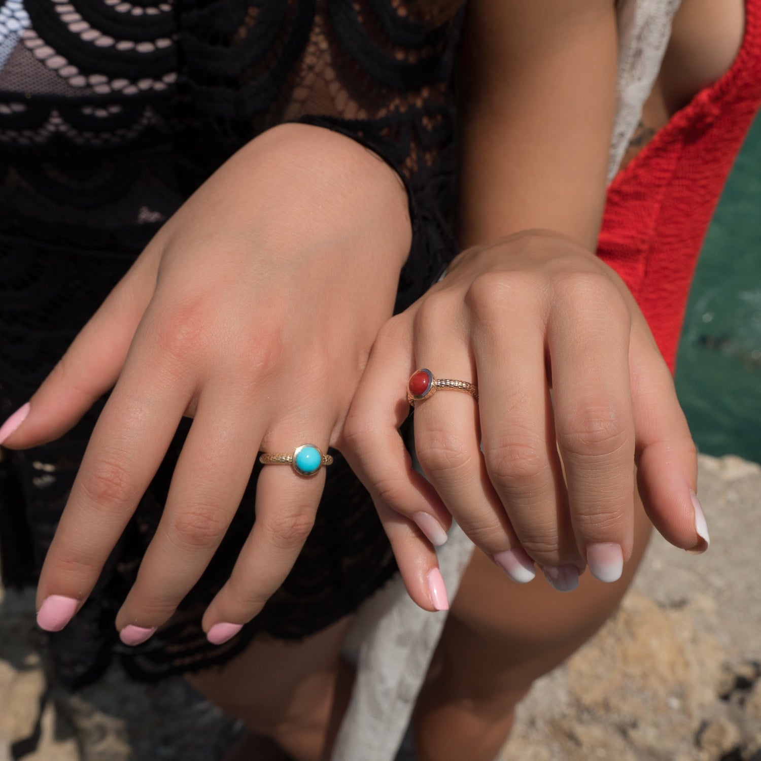 Close-up of two women’s hands wearing turquoise and red coral rings by Jewelry Design House outdoors.