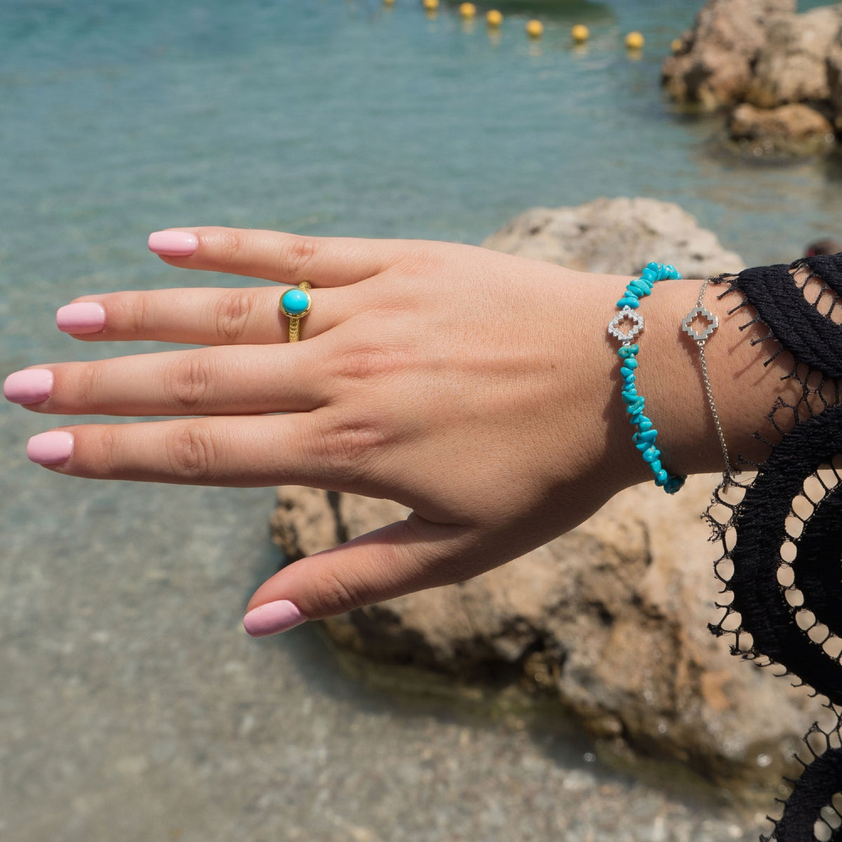Close-up of a woman wearing a turquoise ring, a turquoise beaded bracelet, and a delicate chain bracelet by Jewelry Design House beside seaside rocks.
